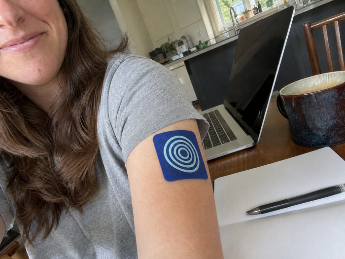 Woman wearing a Pure Patch Focus Patch on her upper arm beside a laptop and open notebook at a kitchen table.