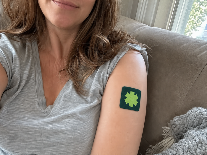 Woman sitting on a sofa by a window with a Pure Patch Recovery Patch on her upper arm.