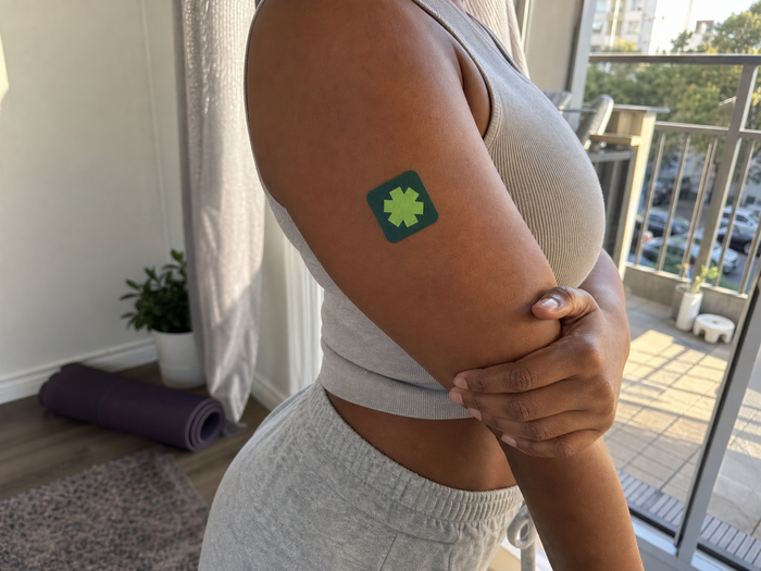 Woman in gray activewear wearing a Pure Patch Recovery Patch on her upper arm beside a balcony door.