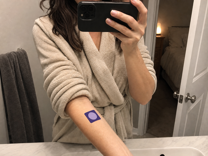 Woman in a bathrobe taking a mirror photo with a Pure Patch Sleep Patch on her forearm.