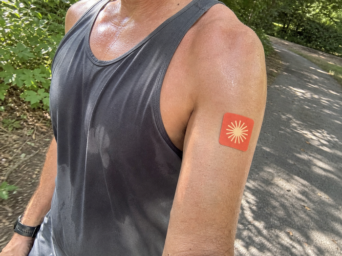 Person in a gray tank top wearing a Pure Patch Energy Patch on their upper arm beside a tree-lined path.