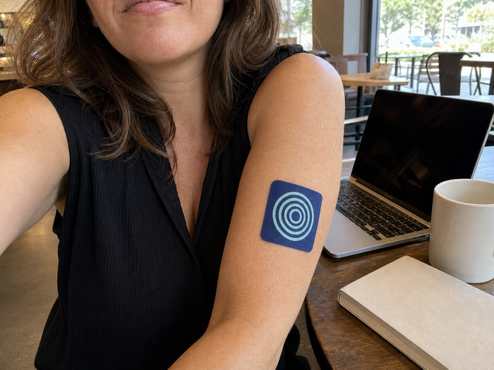 Woman wearing a Pure Patch Focus Patch on her upper arm beside a laptop and mug in a cafe.