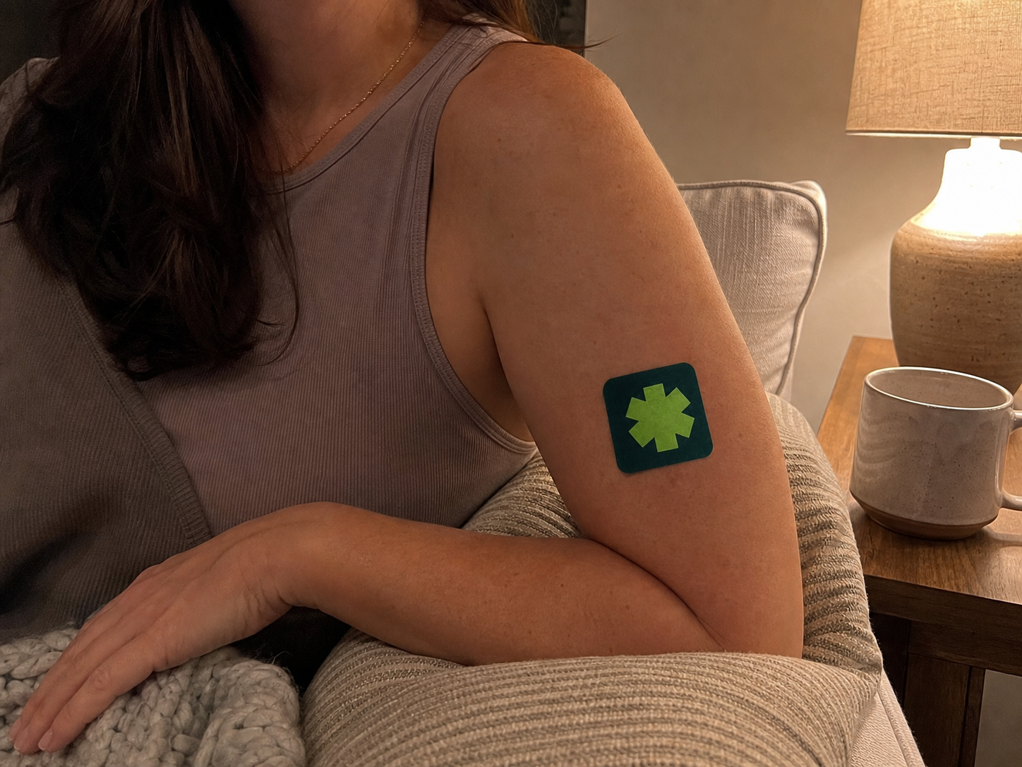 Woman sitting on a sofa wearing a Pure Patch Recovery Patch on her upper arm.